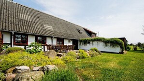 A traditional house with a sloped roof, a front porch with wooden chairs, and a garden with rocks and greenery. - Nydamsgaard "hus lille rede" (Faaborg \/ Bojden)