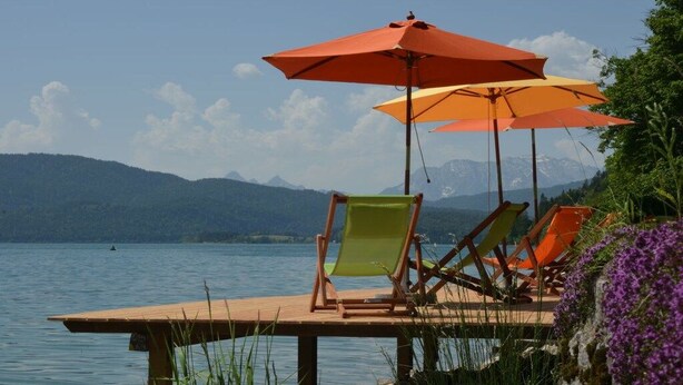 Ein Seeufer mit Stühlen und Sonnenschirmen, mit Blick auf einen ruhigen See und Berge.