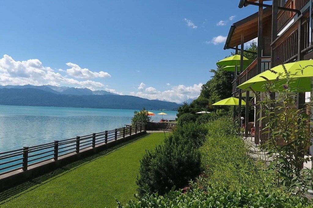 A lakeside view with a wooden deck, umbrellas, and a building with balconies.