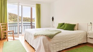 Ein Schlafzimmer mit Bett, Stuhl, Seeblick und Balkon mit grünen Vorhängen.