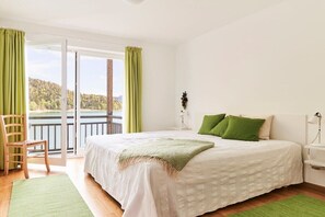 Ein Schlafzimmer mit Bett, Stuhl, Seeblick und Balkon mit grünen Vorhängen.