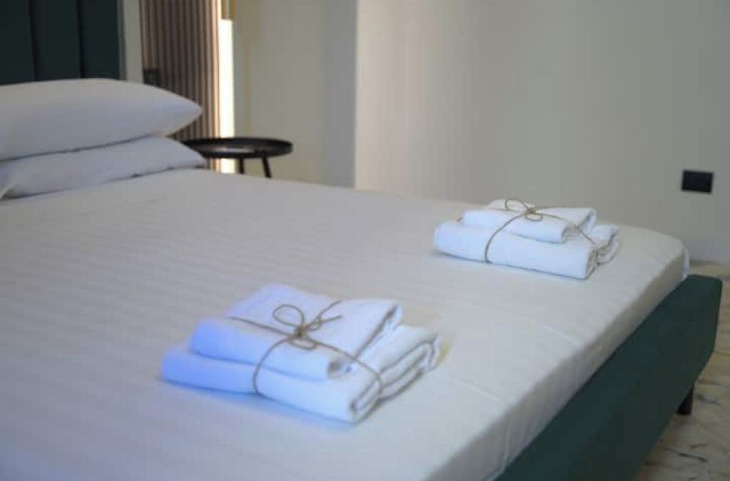 A bed with white linens and towels, a small round table, and a green upholstered bench.