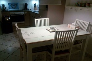 A dining area with a white table and chairs, a kitchen in the background, and a shelf with items. - Small holiday apartment Pappelhof (Fehmarn)