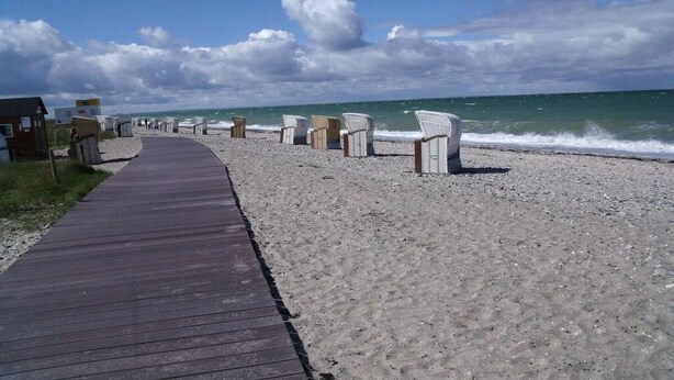 En träpromenad som leder till strandstolar och en liten byggnad på en sandstrand.