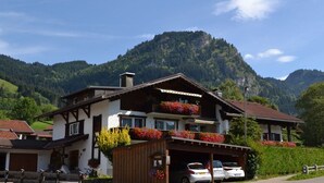 Um edifício de dois andares com varanda, garagem com carros e montanhas ao fundo.