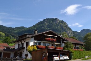 A two-story building with a balcony, a garage with cars, and a mountainous backdrop. - at the E. Schmid guesthouse (Bad Hindelang)