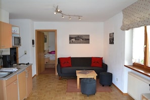 A compact living space with a kitchenette, a sofa with red cushions, a small table, and a window with a view. - at the E. Schmid guesthouse (Bad Hindelang)