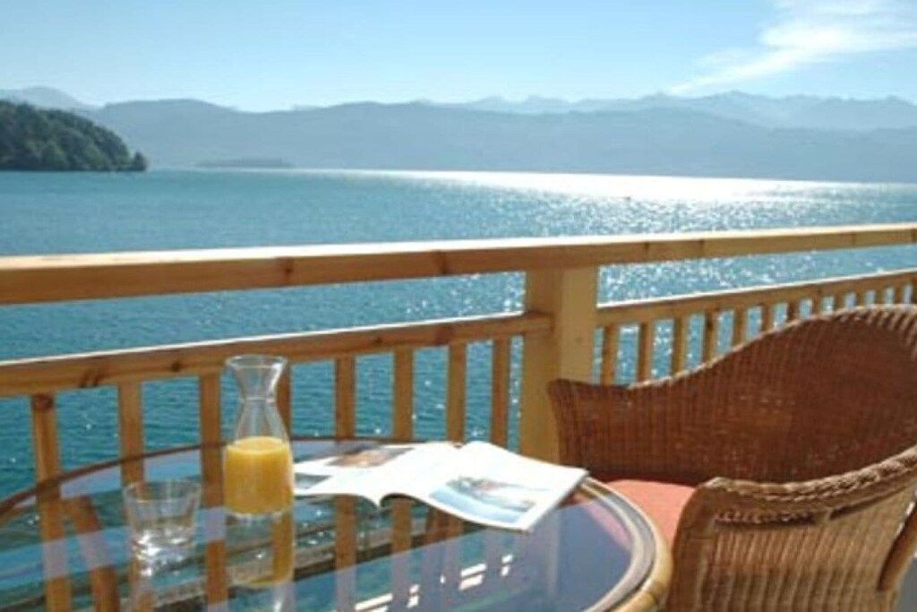 Una terraza con una mesa de vidrio, una silla de mimbre y vista a un lago y montañas.