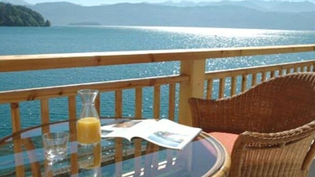 Eine Terrasse mit einem Glastisch, einem Korbstuhl und Blick auf einen See und Berge.