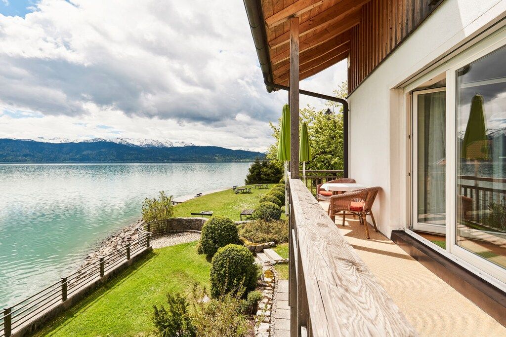 A balcony with a view of a lake and mountains, a wooden deck, and a glass door leading to an outdoor seating area.