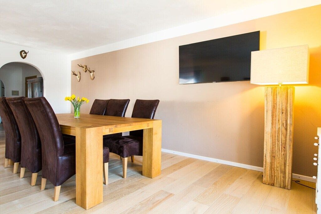 Un coin repas avec une table et des chaises en bois, un vase avec des fleurs, un téléviseur fixé au mur et une lampe.