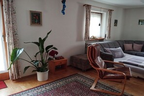 Ein Wohnzimmer mit einer Couch, einem Sessel, einem Topfpflanzen, einem Teppich und einem Fenster mit Vorhängen.