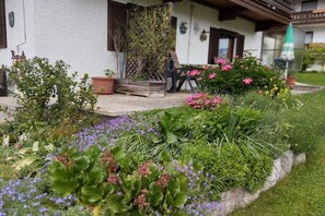 Ein Garten mit verschiedenen Pflanzen und Blumen vor einem Gebäude mit weißer Fassade und Holzläden.