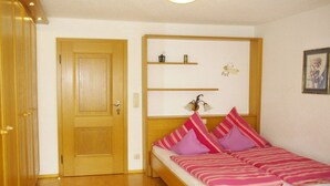 Quarto com porta de madeira, cama com roupa de cama rosa e roxa, e uma prateleira com objetos decorativos.