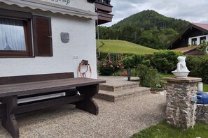 A rustic building with a wooden bench outside, a stone wall, and a view of a green hill. - Haus Berggruß EG with Chiemgau map (Ruhpolding)