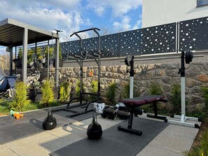Une salle de sport en plein air équipée de divers appareils, un espace de repos couvert et un mur en pierre servant de décor.