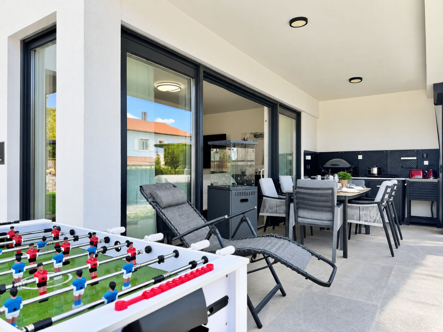 A modern outdoor patio with a foosball table, a dining area with chairs and a table, and a kitchen area with appliances.