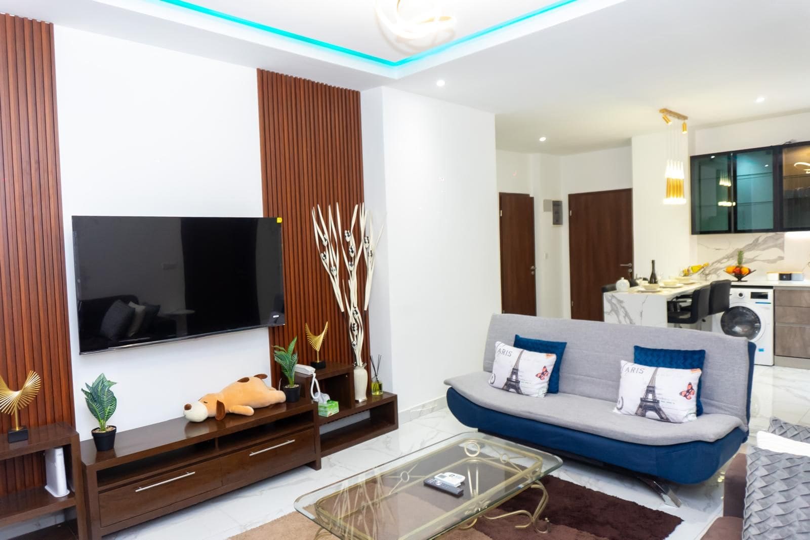 Una sala moderna con un televisor de pantalla plana, un sofá con cojines decorativos, una mesa de centro y una cocina al fondo.