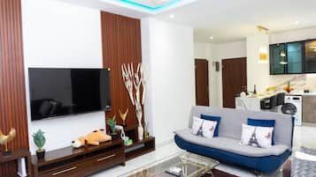 Una sala moderna con un televisor de pantalla plana, un sofá con cojines decorativos, una mesa de centro y una cocina al fondo.