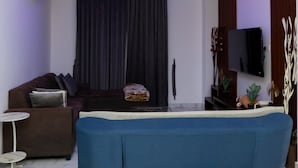 A modern living room with a blue sofa, a flat-screen TV, a ceiling light fixture, and a dark curtain. - Peravi at The Essence  (Accra)