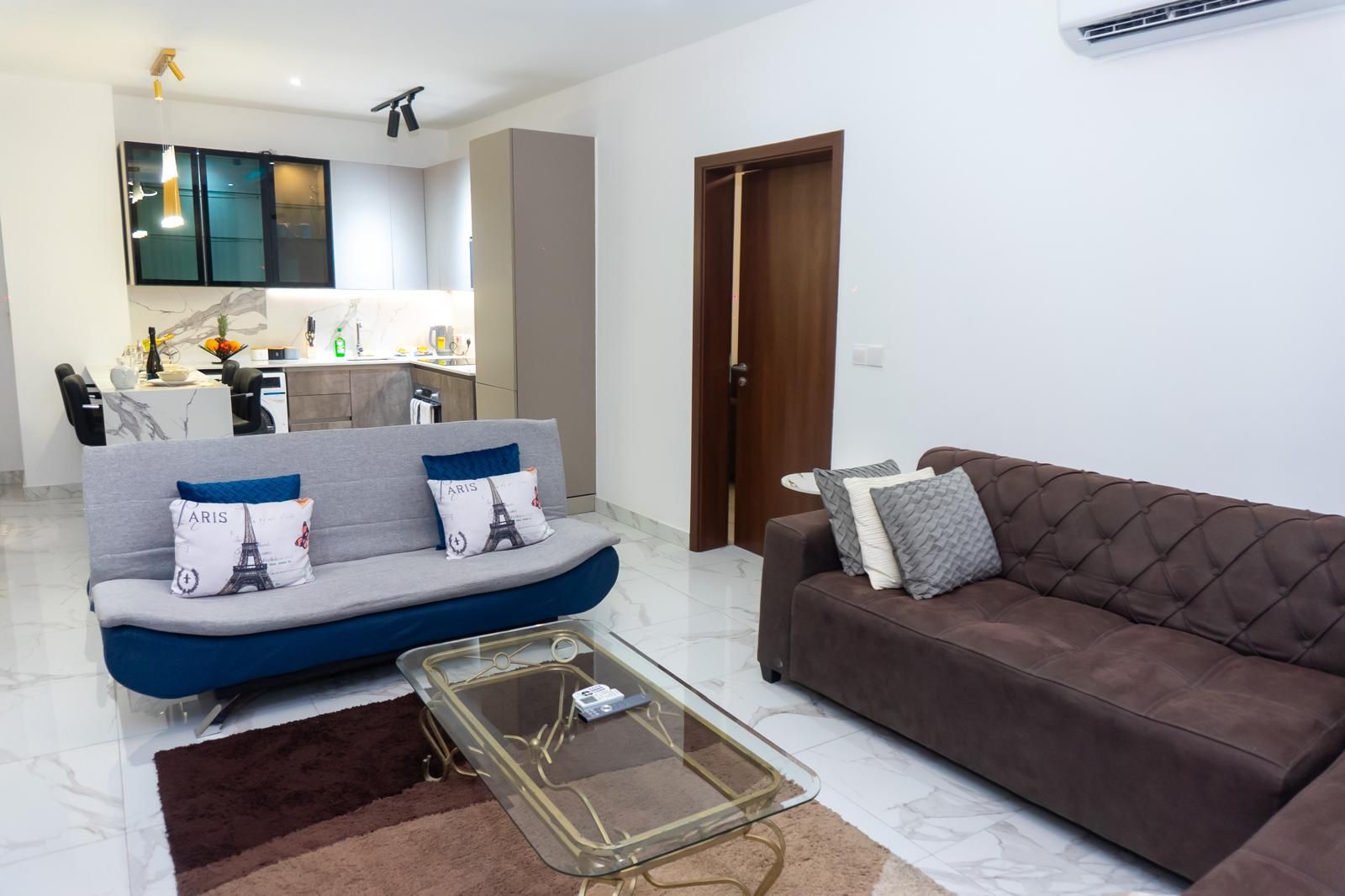 Ein Wohnzimmer mit einer Couch, einem braunen Sofa, einem Glastisch und einer Küche im Hintergrund.