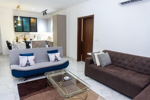 Sala de estar com sofá, um sofá marrom, uma mesa de centro de vidro e uma cozinha ao fundo.