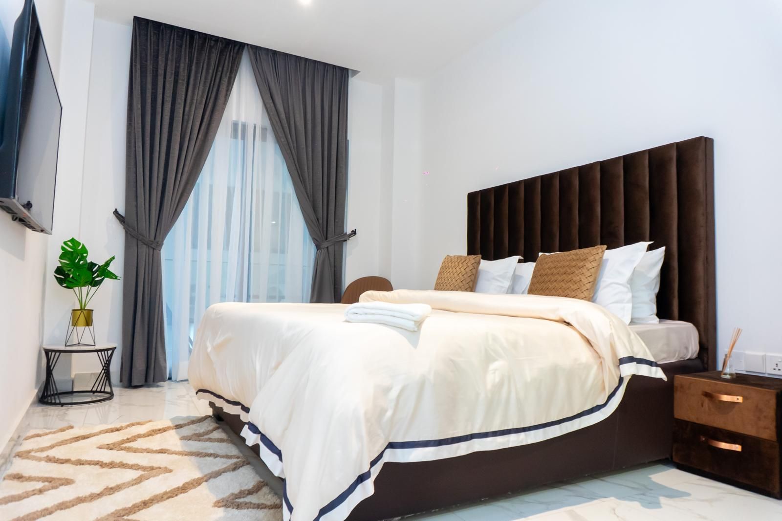 Ein modernes Hotelzimmer mit einem großen Bett, einem Nachttisch, einem kleinen Beistelltisch, einer Zimmerpflanze und Blick auf die Stadt durch das Fenster.