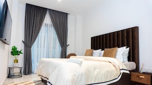A modern hotel room with a large bed, a bedside table, a small side table, a potted plant, and a view of the city through the window. - Peravi at The Essence  (Accra)