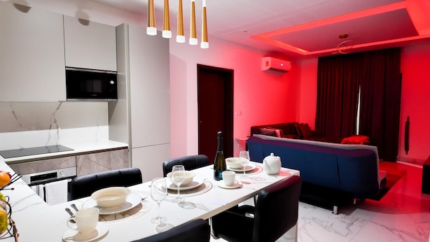 A modern dining area with a white table set for four, a kitchen with a built-in microwave, and a red-lit living area with a sofa and curtains. - Peravi at The Essence  (Accra)