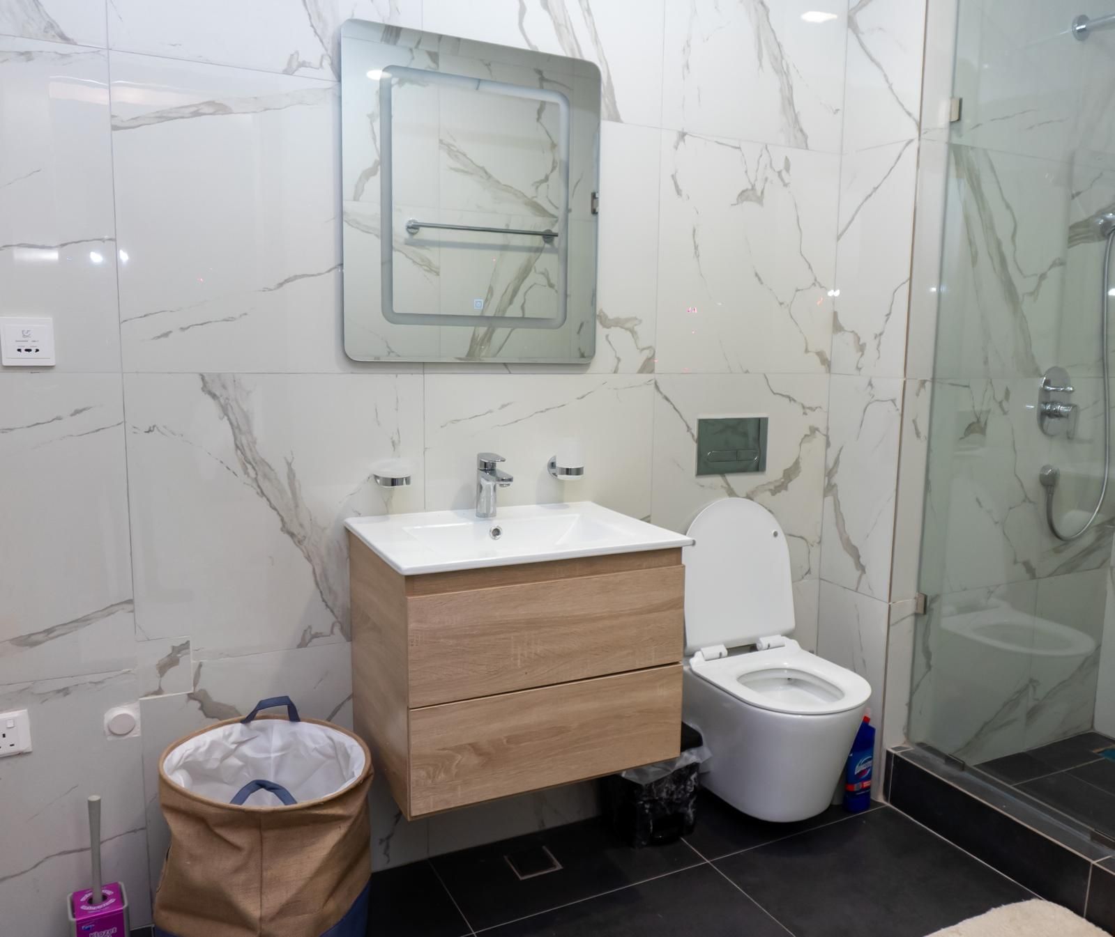 A bathroom with a marble wall, a wooden vanity, a white sink, a toilet, and a glass shower enclosure.