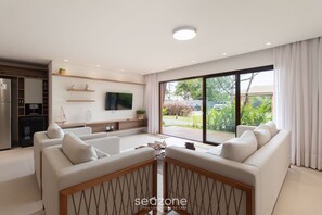 A modern living room with a sofa, TV, and a view of the outdoors. - House in Arraial D'ajuda With Pool Pcr0002 (Porto Seguro)