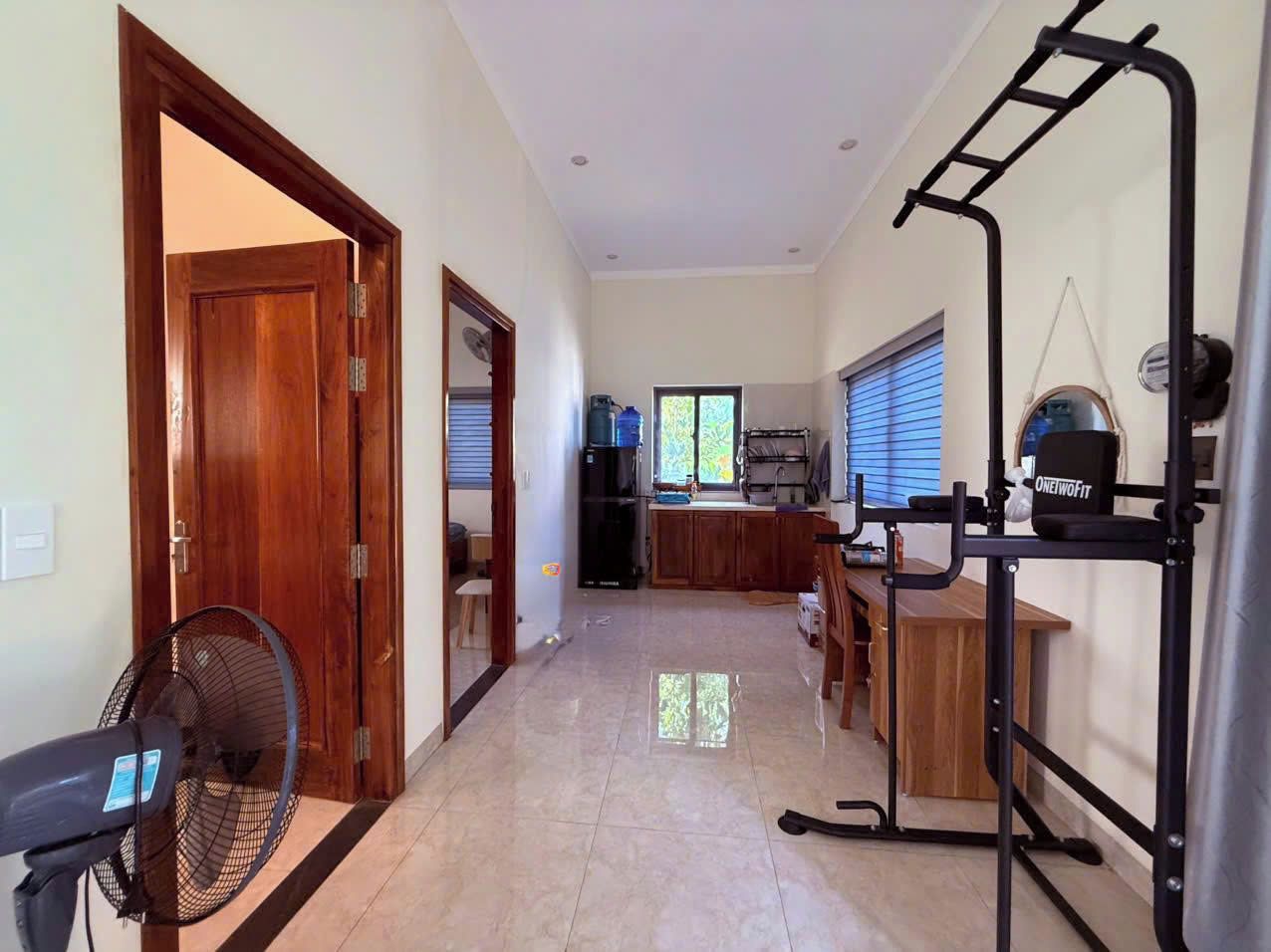 A spacious living area with a wooden door, a fan, a workout station, and a kitchen in the background.