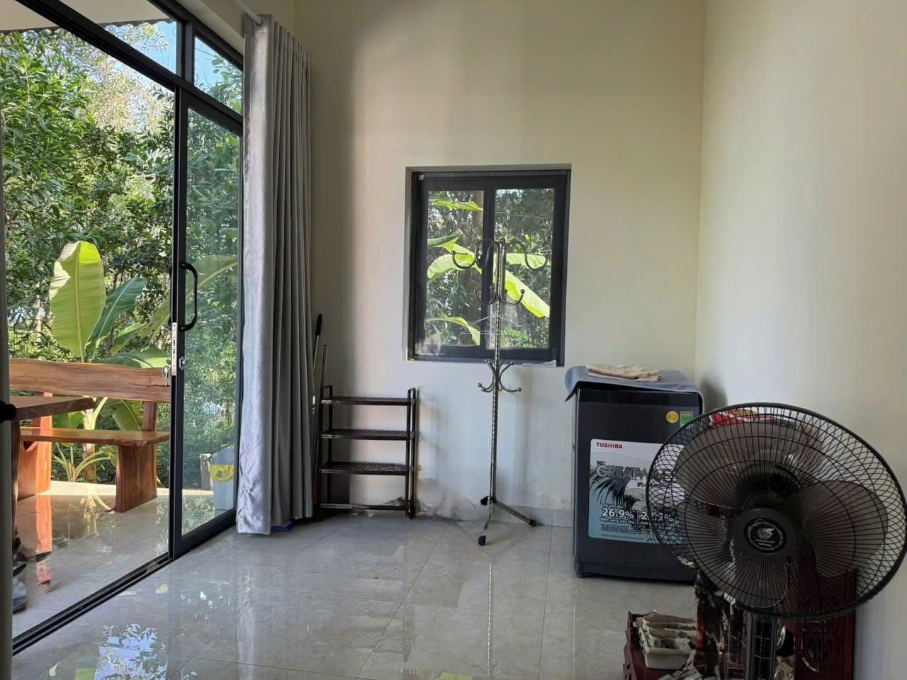 A room with a sliding glass door leading to a balcony, a standing fan, and a window with a view of greenery.
