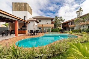 A modern outdoor pool area with a barbecue setup, surrounded by lush greenery and a wooden deck. - House in Arraial D'ajuda With Pool Pcr0009 (Porto Seguro)