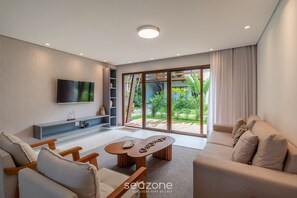 A modern living room with a sofa, coffee table, TV, and sliding glass doors leading to a garden. - House in Arraial D'ajuda With Pool Pcr0009 (Porto Seguro)