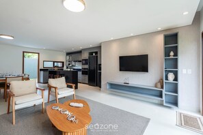 Una sala de estar moderna con una mesa de centro de madera, un televisor de pantalla plana montado en la pared y una cocina integrada con refrigerador y microondas.