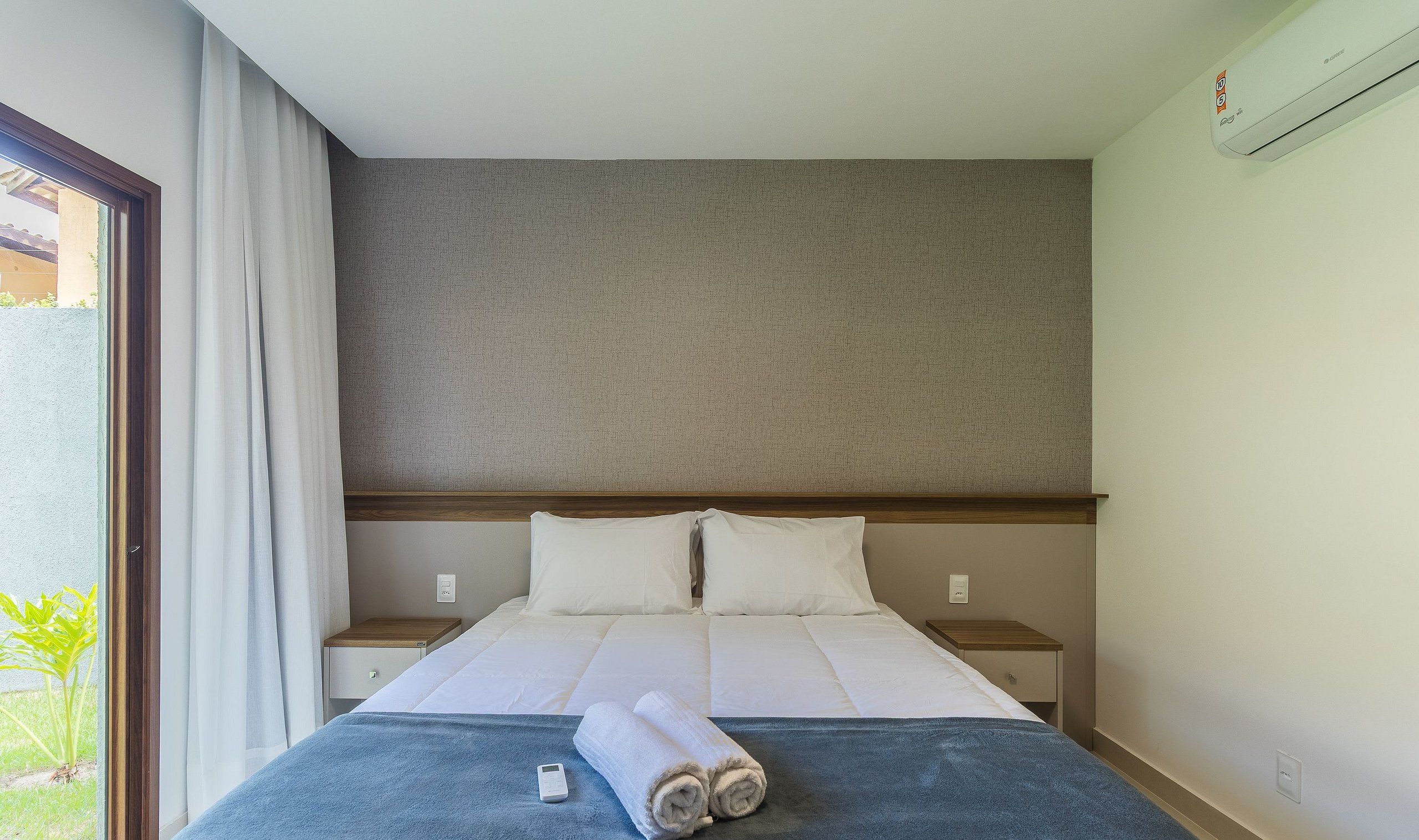 A hotel room with a bed, a nightstand, a wall-mounted air conditioner, and a window with curtains.