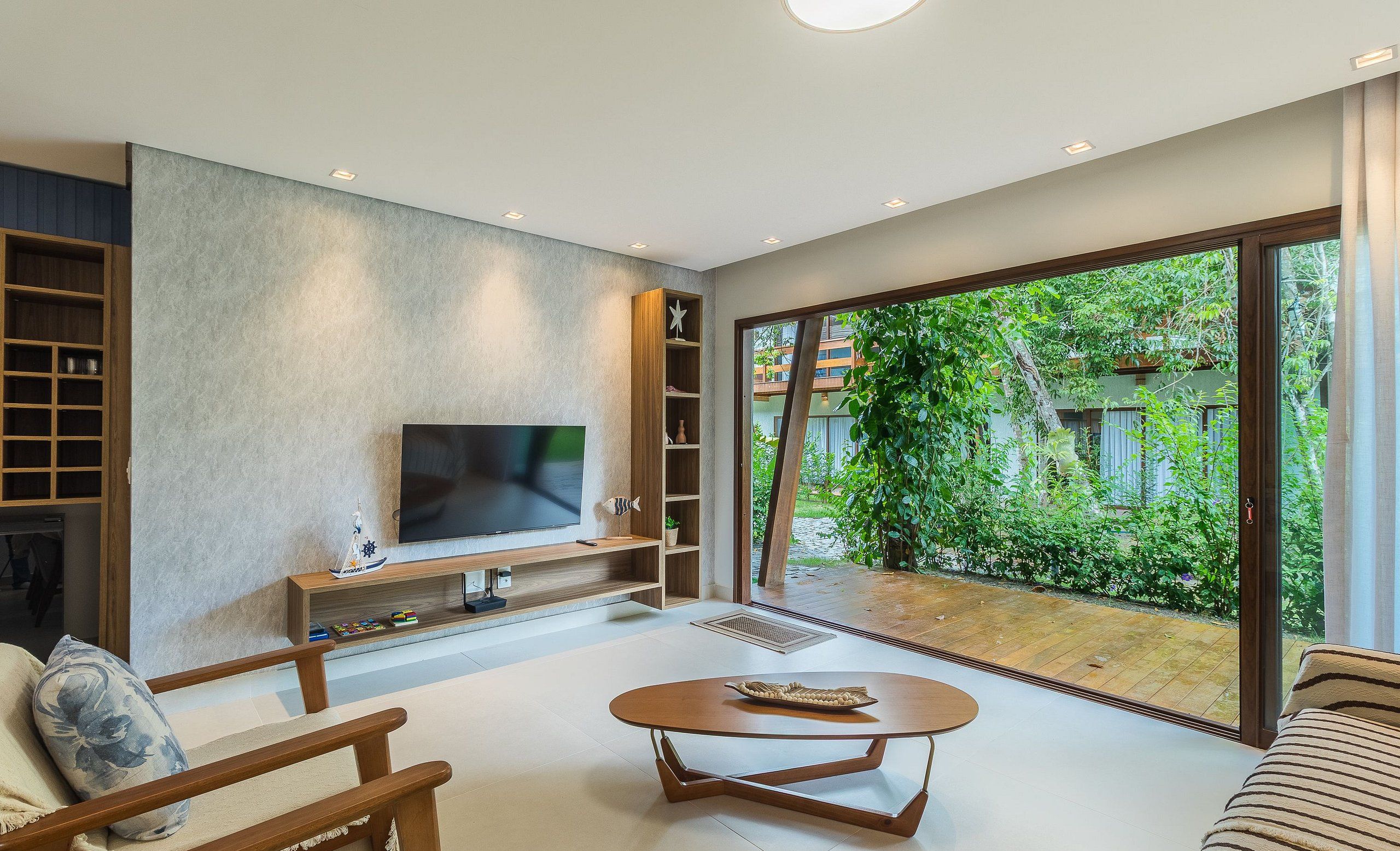 A modern living room with a large TV, wooden shelves, and a view of greenery through large windows.