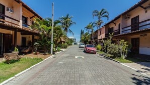 A residential street with houses, palm trees, and a red car parked on the side. - House in Cond. w/ Pool - Cabo Frio Vdp0029 (Cabo Frio)