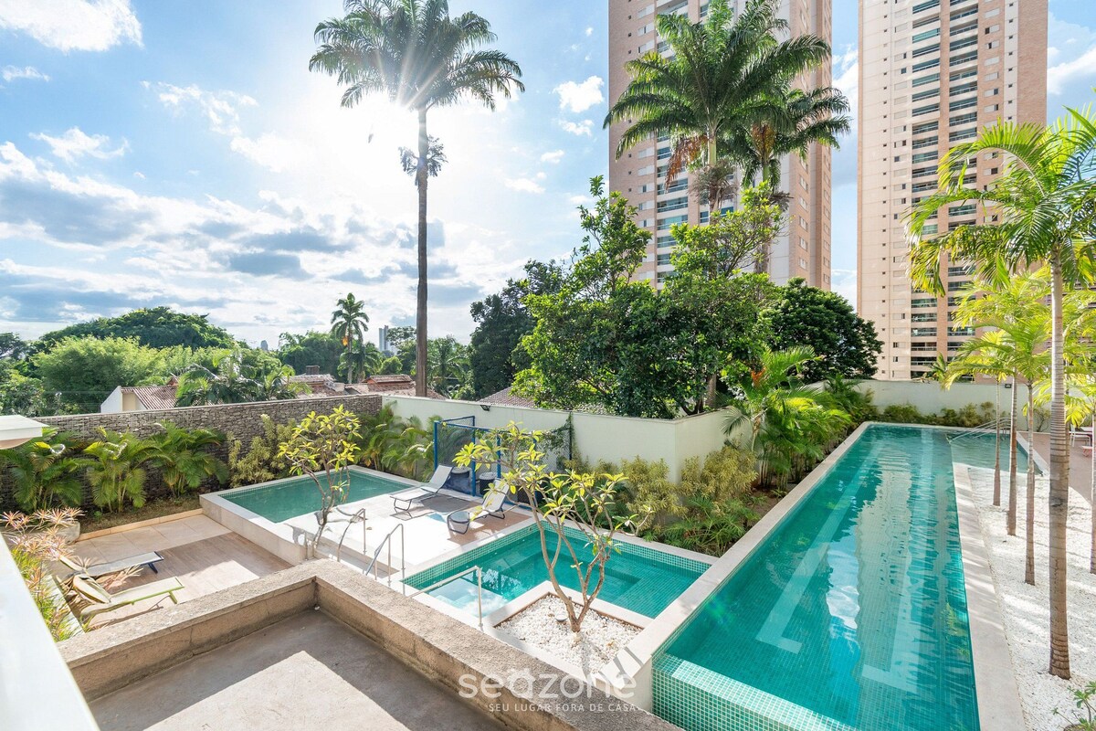 A rooftop swimming pool with lounge chairs, surrounded by greenery and tall buildings.