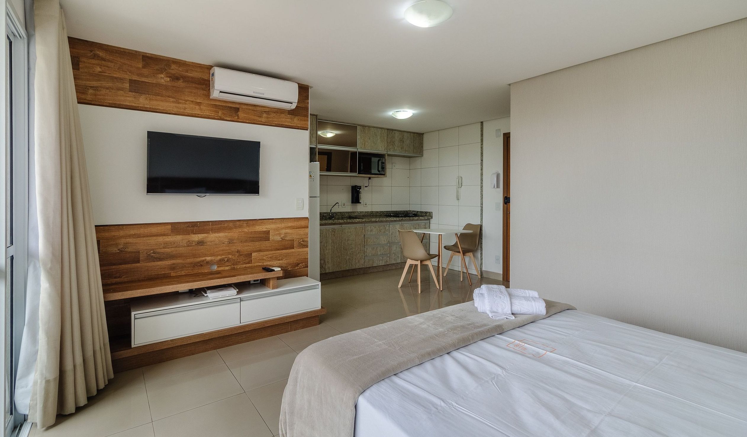 A hotel room with a bed, a TV mounted on a wooden wall, a kitchenette, and a dining area with chairs.