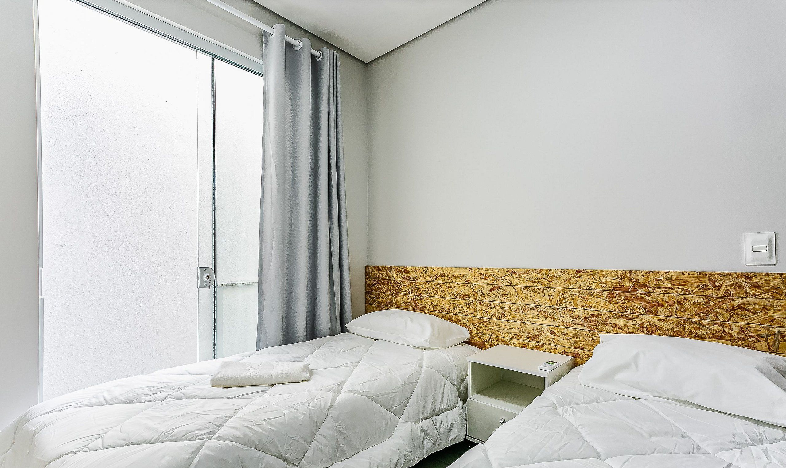 A hotel room with two beds, a window with curtains, a wooden headboard, and a small bedside table.