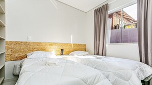 Quarto com cama, cabeceira, janela com cortinas e vista para prédios.