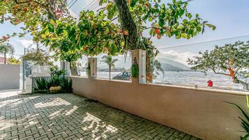 A paved outdoor area with a view of the sea, a wall with a window, and some greenery.