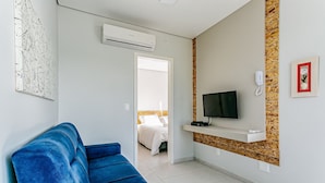 A hotel room with a blue sofa, a TV on a wooden shelf, a painting, and an air conditioning unit. - Fit w/ Beautiful View of Lagoa da Conceicao Sdr002 (Florianópolis)