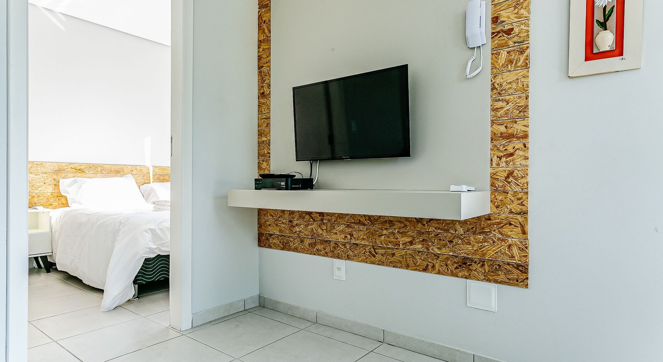 Une chambre d’hôtel avec un téléviseur fixé au mur, une étagère blanche en dessous et un lit avec une literie blanche.