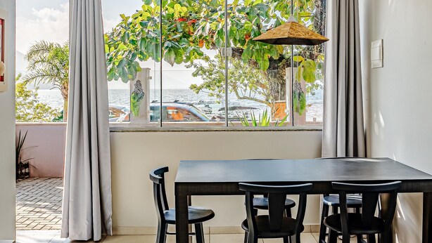 Uma área de refeições com mesa e cadeiras, vista para o mar através de amplas janelas e uma planta pendente.