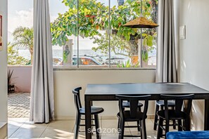 Ein Essbereich mit Tisch und Stühlen, Blick auf das Meer durch große Fenster und eine hängende Pflanze.