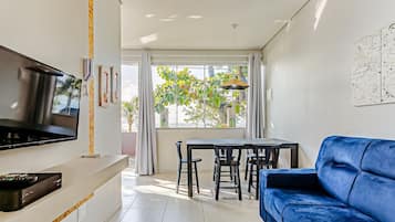 Una sala moderna con un televisor de pantalla plana, un sofá azul, una mesa de comedor con sillas y un ventanal con cortinas.
