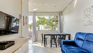 En moderne stue med et fladskærms-tv, en blå sofa, et spisebord med stole og et stort vindue med gardiner.